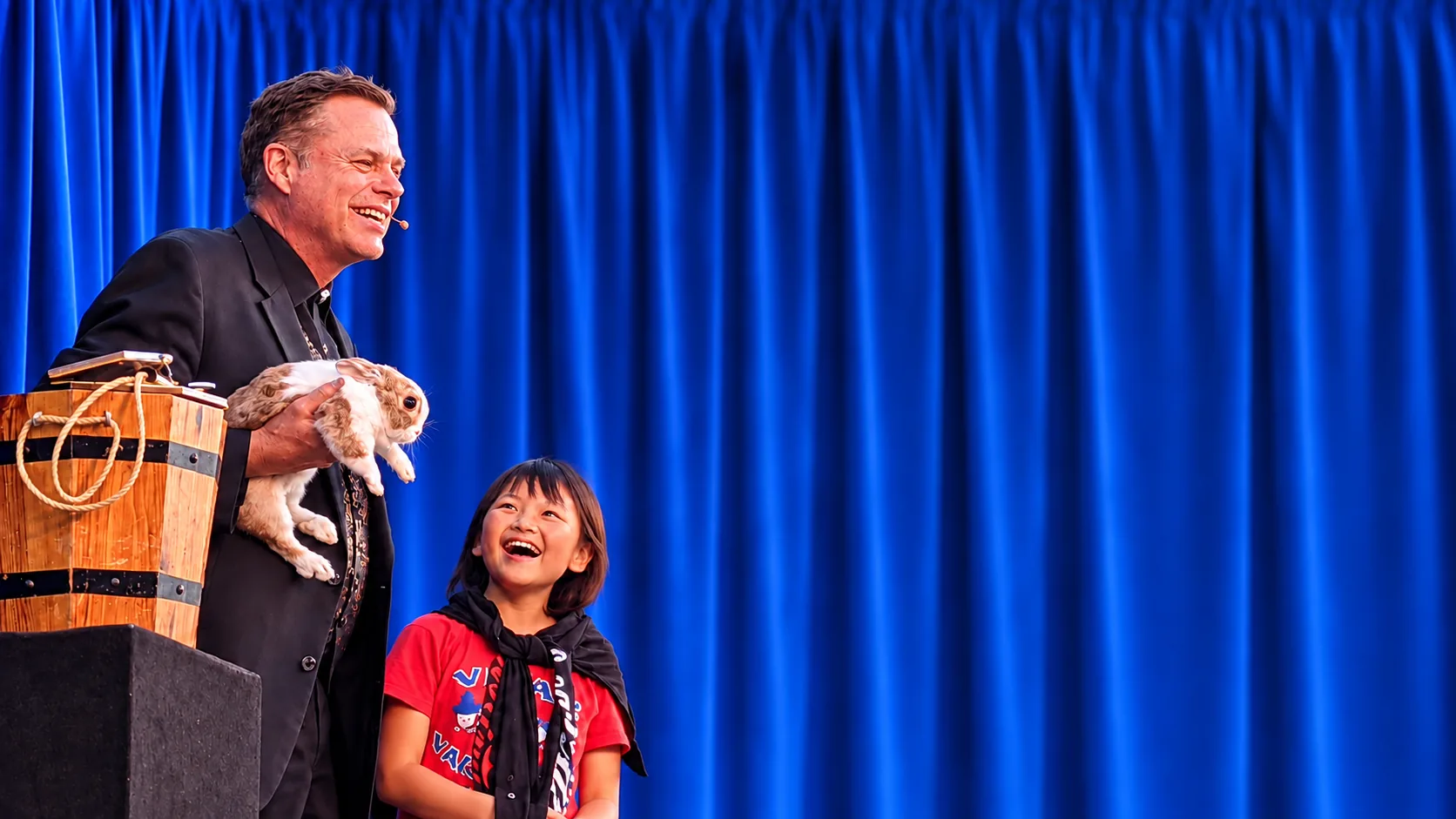 Brock Edwards holding a live rabbit while a delighted elementary student reacts during The Magic of Literature reading assembly